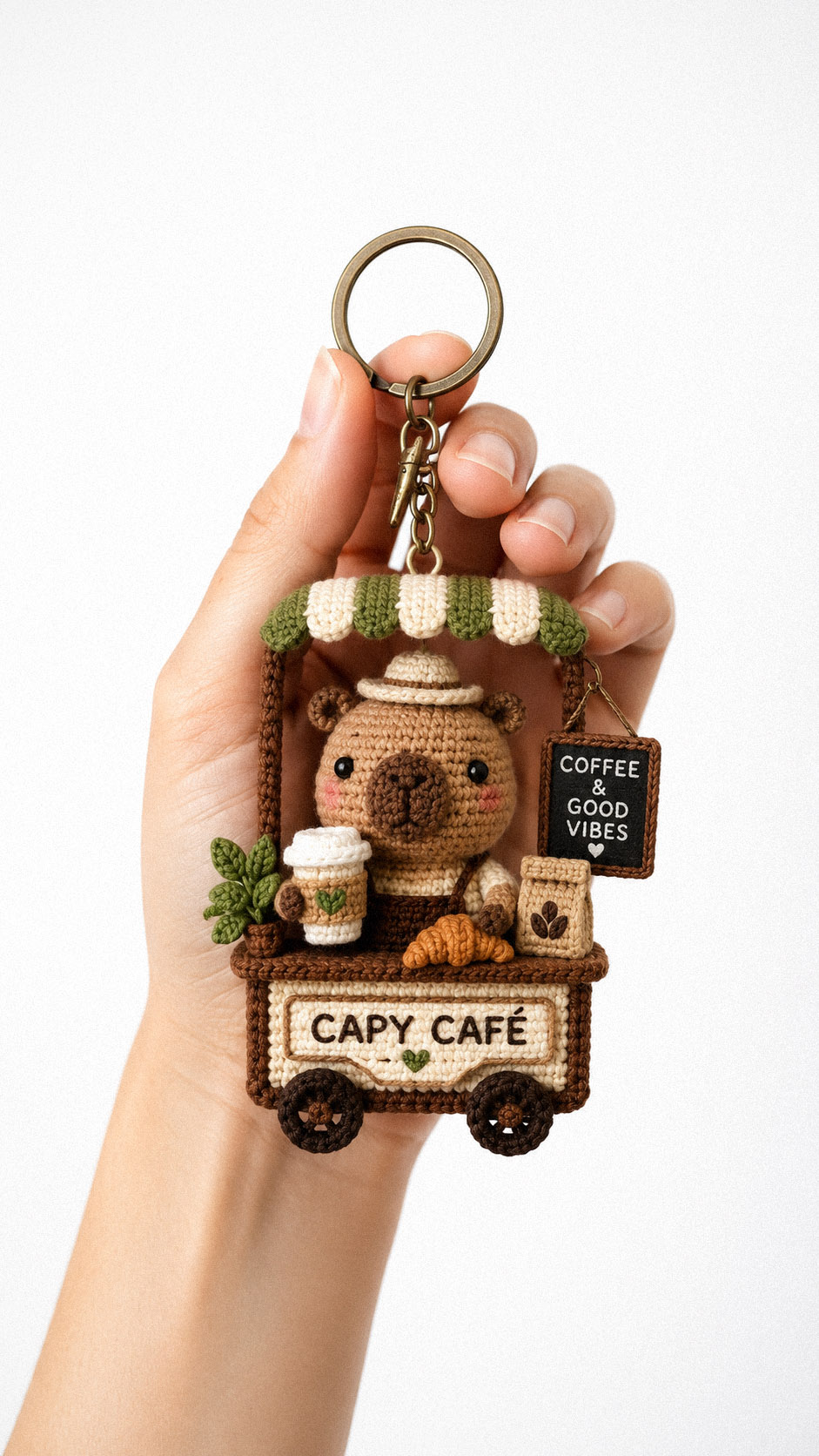Crochet capybara barista keychain with a coffee cup and croissant charm held in a hand on a clean white background