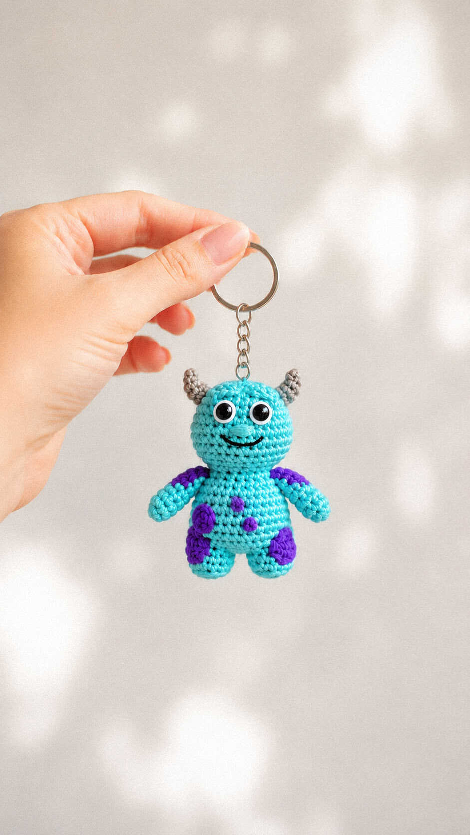 Tiny blue crochet monster keychain with playful purple spots and small horns on a soft neutral background